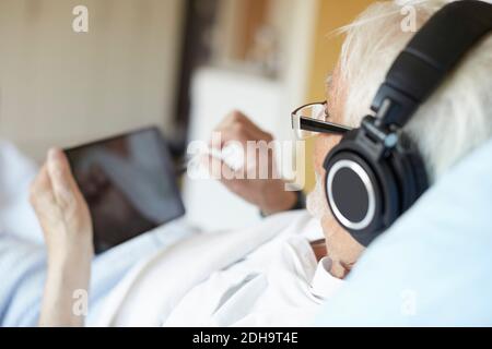 Vista sulla spalla di un uomo anziano che indossa le cuffie mentre utilizzo di un tablet digitale in ospedale Foto Stock