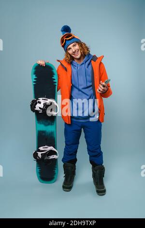 Bel sportivo felice in posa con cellulare e snowboard isolato sopra sfondo blu Foto Stock