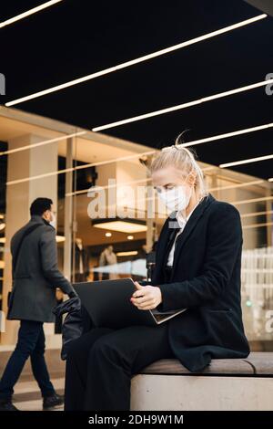 Donna d'affari con maschera seduta con laptop all'aperto Foto Stock