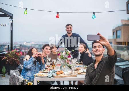 Uomo sorridente che prende selfie attraverso lo smartphone mentre ci si diverte amici in riunione sociale sulla terrazza dell'edificio Foto Stock