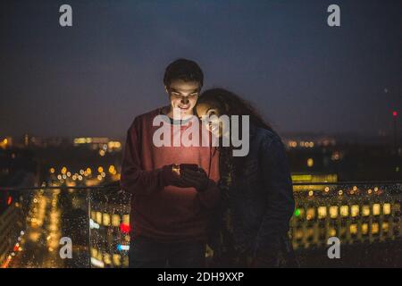 Giovane uomo che mostra smartphone a un'amica durante il social riunione sulla terrazza di notte Foto Stock