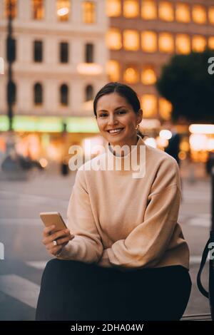 Ritratto di una giovane donna sorridente che usa il telefono mentre si è seduti città Foto Stock