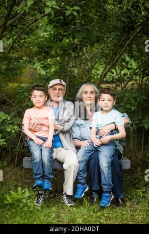 Ritratto di nonni seduti con due nipoti su panchina contro piante nel cortile posteriore Foto Stock