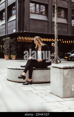 Donna d'affari con cuffie intrauricolari che guardano lontano mentre si siede all'aperto Foto Stock