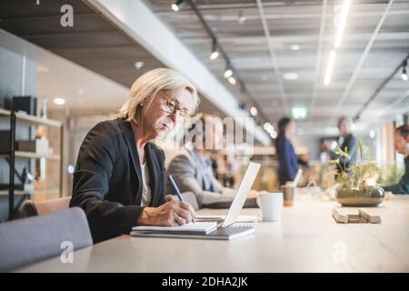L'imprenditore femminile anziano scrive nel diario mentre lavora al tavolo in ufficio Foto Stock