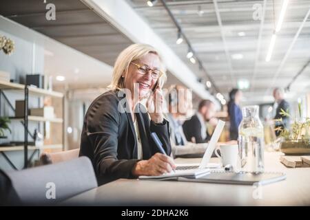 Donna d'affari sorridente che parla sul telefono cellulare e scrive nel diario mentre si lavora alla scrivania dell'ufficio Foto Stock