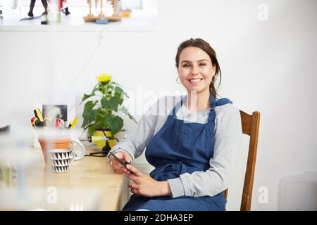 Ritratto di sorridente giovane ingegnere con smartphone seduto al tavolo in officina Foto Stock