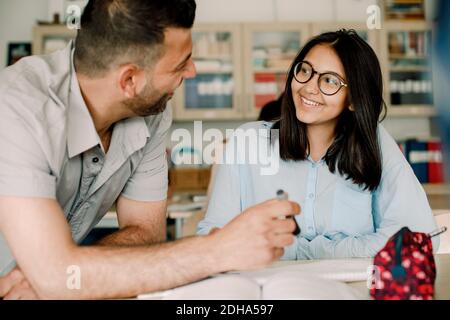 Sorridente insegnante di sesso maschile che spiega lo studente mentre si appoggia al tavolo in aula Foto Stock