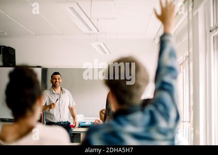 Studente maschile con mano sollevata mentre sorride tutor in piedi in aula Foto Stock