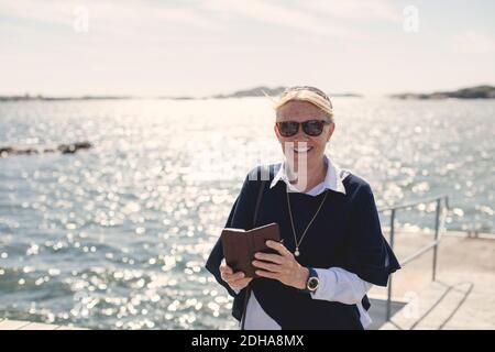 Ritratto di donna anziana sorridente che indossa occhiali da sole mentre si tiene in movimento telefono contro lago Foto Stock