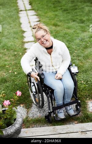 Ritratto a lunghezza intera di una donna sorridente e disabile seduta su sedia a rotelle in cortile Foto Stock