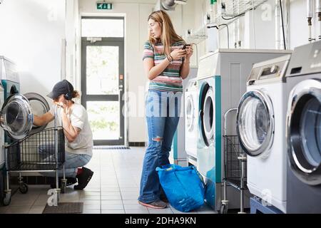 Donna che usa il telefono cellulare mentre l'uomo fa il bucato alla lavanderia automatica Foto Stock