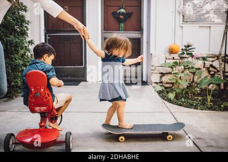 Figlia skateboarding con l'aiuto di madre mentre il figlio è seduto sopra triciclo Foto Stock