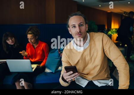 Ritratto di uomo d'affari sicuro che tiene lo smartphone mentre si siede contro colleghi di lavoro in caffetteria Foto Stock