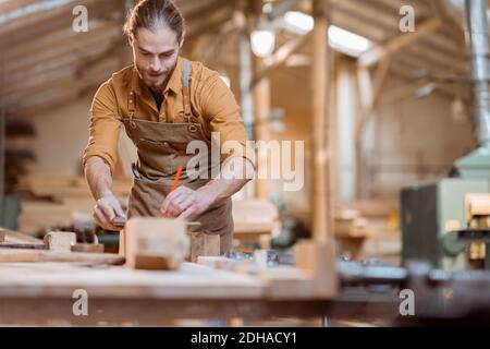 Bel falegname che lavora con un legno, segnando una tavola con una matita nel laboratorio di Falegnameria Foto Stock