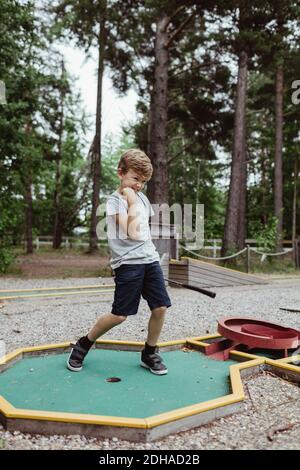 Lunghezza completa di ragazzo allegro che gioca a minigolf in cortile durante le vacanze Foto Stock