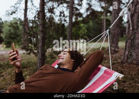 Sorridendo un giovane uomo utilizzando il telefono cellulare mentre si rilassa in amaca contro gli alberi Foto Stock