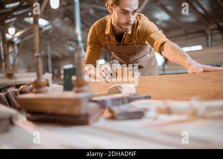 Bel falegname che lavora con un legno, guardando la barra, controllando la qualità in officina Foto Stock