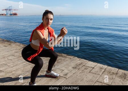 Tiro esterno di workout attraente donna di idoneità vicino al mare, facendo esercizi di squadra e guardando la macchina fotografica Foto Stock