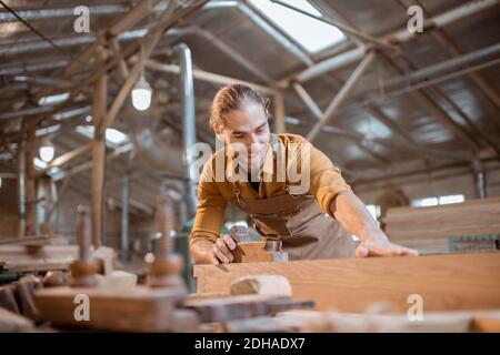 Bel falegname che lavora con un legno, guardando la barra, controllando la qualità in officina Foto Stock