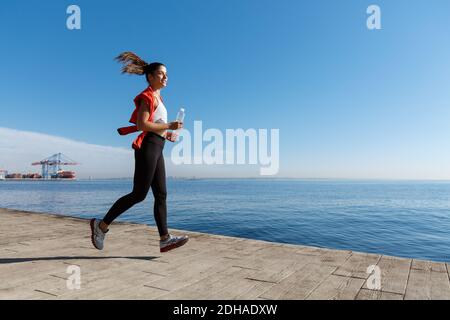 Vista laterale della giovane donna atletica che corre lungo il lungomare. Donna sportiva che fa jogging vicino al mare con una bottiglia d'acqua Foto Stock