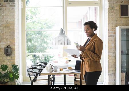 Felice architetto femminile utilizzando il telefono mentre si sta in piedi al tavolo casa Foto Stock