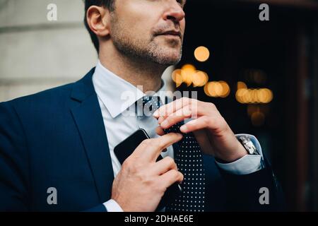 Sezione centrale del businessman che regola la cravatta mentre tiene il telefono cellulare Foto Stock