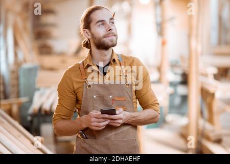 Ritratto di un bel falegname con smartphone, che ha un freno durante la lavorazione del legno in officina Foto Stock