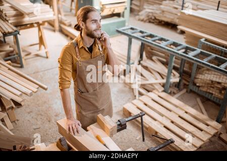 Bel falegname che parla al telefono, con un freno durante il lavoro in legno in officina Foto Stock