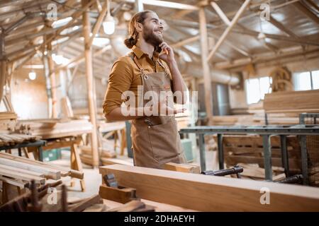 Bel falegname che parla al telefono, con un freno durante il lavoro in legno in officina Foto Stock