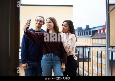 Donna sorridente che prende selfie con i giovani amici mentre si sta in piedi balcone in affitto appartamento Foto Stock