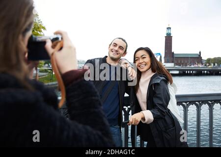 Donna che fotografa amici sorridenti in piedi sul ponte facendo corrimano città contro il cielo limpido Foto Stock