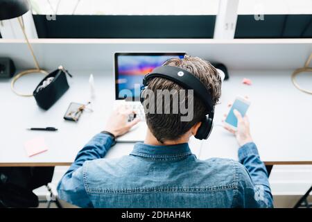 Visualizzazione ad alto angolo di un uomo d'affari che indossa le cuffie utilizzando tecnologie portatili mentre si è seduti alla scrivania dell'ufficio Foto Stock