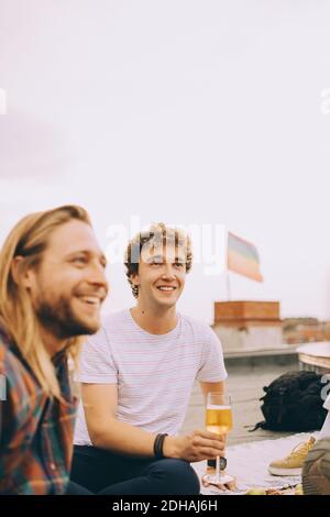 Sorridi gli amici maschili gustando la birra mentre si guarda via alla terrazza durante la festa contro il cielo Foto Stock