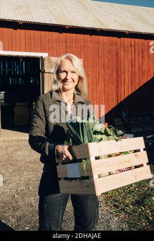 Ritratto di donna matura sorridente con cassa piena di verdure con fienile in background Foto Stock