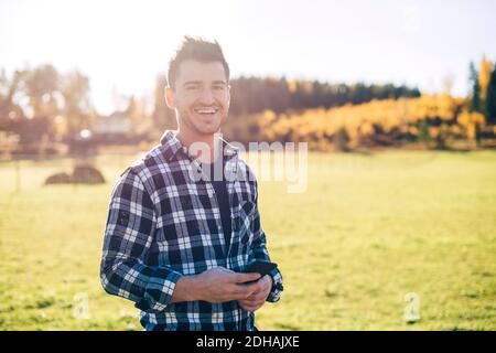 Ritratto di sorridente metà adulto agricoltore maschio che tiene telefono cellulare in piedi sul campo Foto Stock