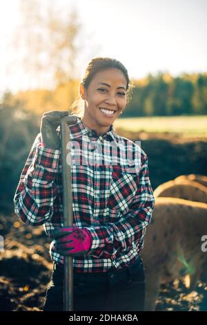 Ritratto di sorridente metà adulta femmina agricoltore che tiene la pala con maiali in background in azienda agricola biologica Foto Stock
