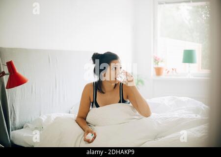 Donna che beve acqua mentre tiene le pillole sul letto a casa Foto Stock