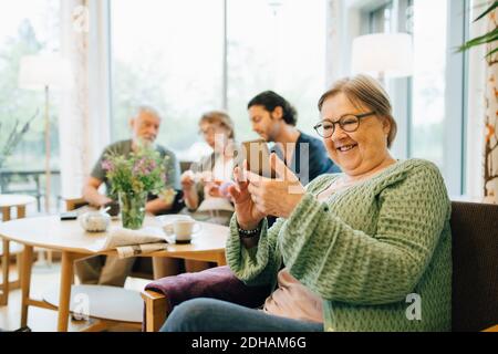 Sorridente donna anziana in pensione che usa lo smartphone mentre si è seduti sedia con amici e caregiver in background Foto Stock