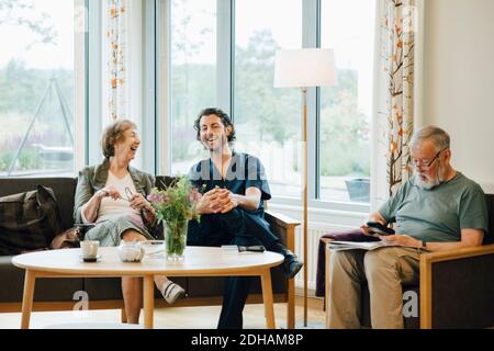 Donna anziana allegra che parla con l'infermiera maschile seduto sul divano contro finestra a casa di cura Foto Stock