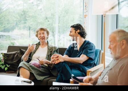 Donna anziana sorridente che parla con l'infermiera maschile seduta sul divano contro finestra a casa di cura Foto Stock