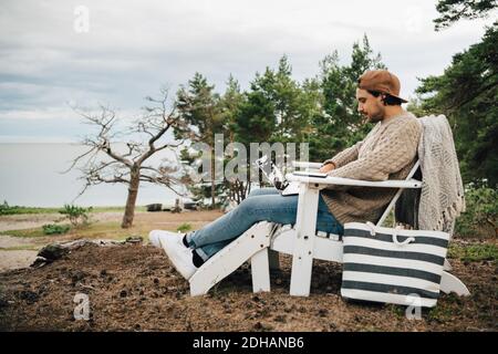 Vista laterale di un uomo che usa un computer portatile mentre si è seduti su Adirondack sedia a foresta Foto Stock