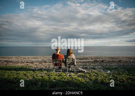 Vista posteriore degli amici seduti sulla sedia in spiaggia contro cielo nuvoloso Foto Stock