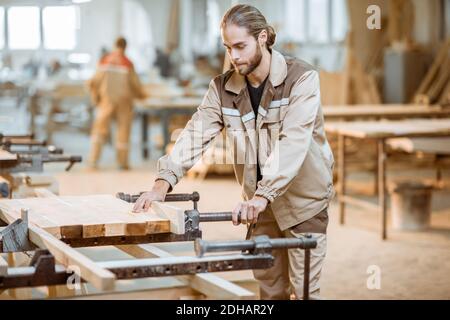 Bel falegname in uniforme incollaggio di barre di legno con pressioni a mano alla produzione di carpenteria Foto Stock