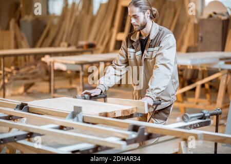 Bel falegname in uniforme incollaggio di barre di legno con pressioni a mano alla produzione di carpenteria Foto Stock