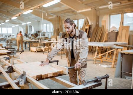 Bel falegname in uniforme incollaggio di barre di legno con pressioni a mano alla produzione di carpenteria Foto Stock