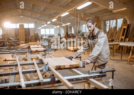 Bel falegname in uniforme incollaggio di barre di legno con pressioni a mano alla produzione di carpenteria Foto Stock