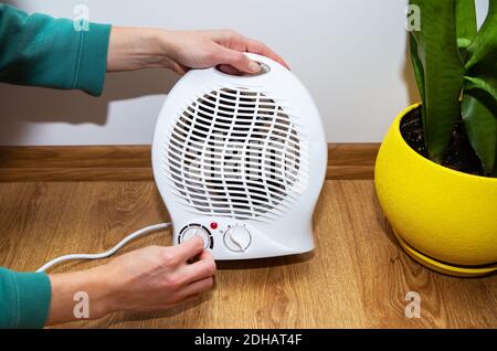 Una ragazza tiene le mani vicino a un riscaldatore di plastica del ventilatore e le riscalda le mani, regolando la temperatura a casa, primo piano Foto Stock