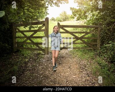 Tutta la lunghezza della ragazza sorridente in piedi da cancello di legno sopra sentiero tra piante Foto Stock