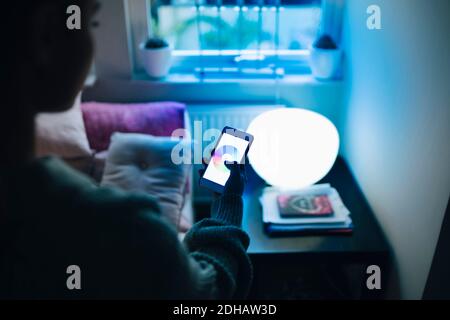 Immagine ritagliata di una ragazza adolescente che usa lo smartphone mentre si è in piedi da letto illuminato in camera Foto Stock